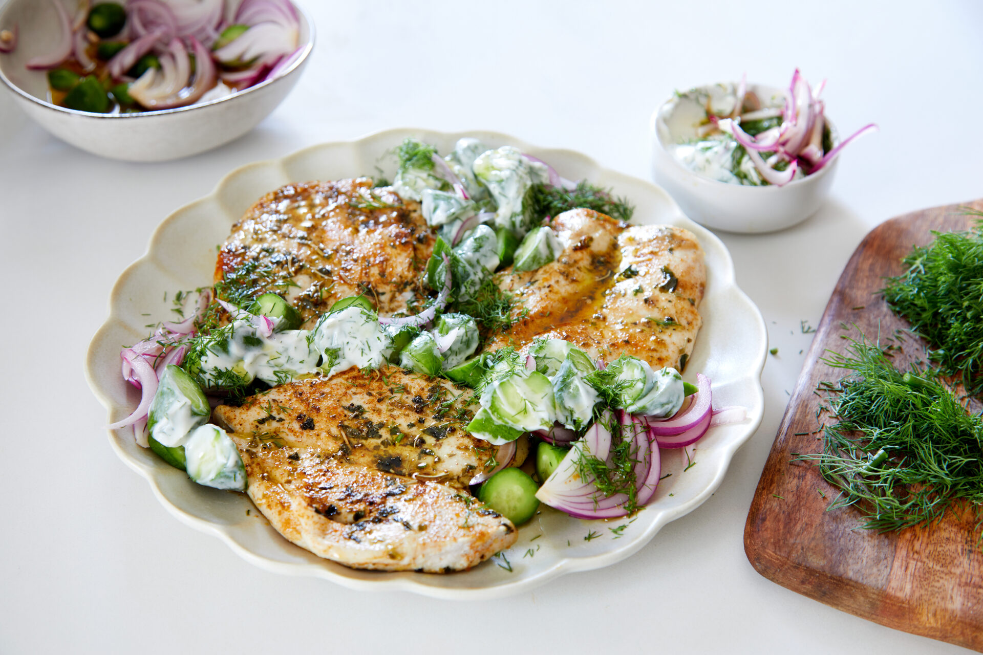 Este prezentată o farfurie cu piept de pui la grătar și salată de castraveți cu iaurt, garnisită cu ceapă feliată și ierburi aromatice, alături de boluri mici cu ingrediente suplimentare pentru salată.