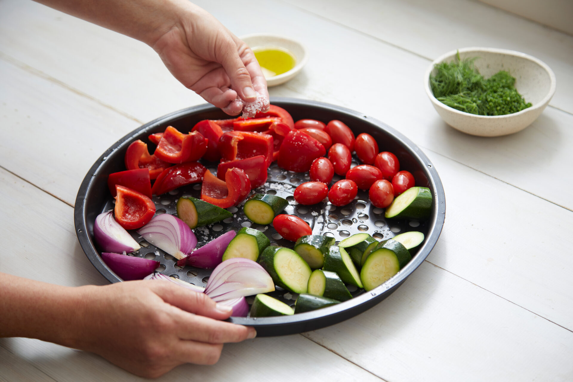 Se așează legume mediteraneene coapte cu ierburi și brânză feta în coșul friteuzei cu aer cald, inclusiv ardei, roșii, dovlecei și ceapă, având lângă ele boluri mici cu ulei și ierburi tocate.